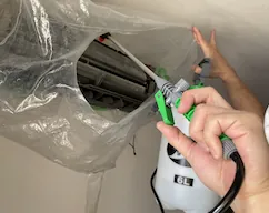 合同会社JAOのスタッフが、洗浄カバーを付けたエアコンに洗剤を吹き付けている実際の作業写真